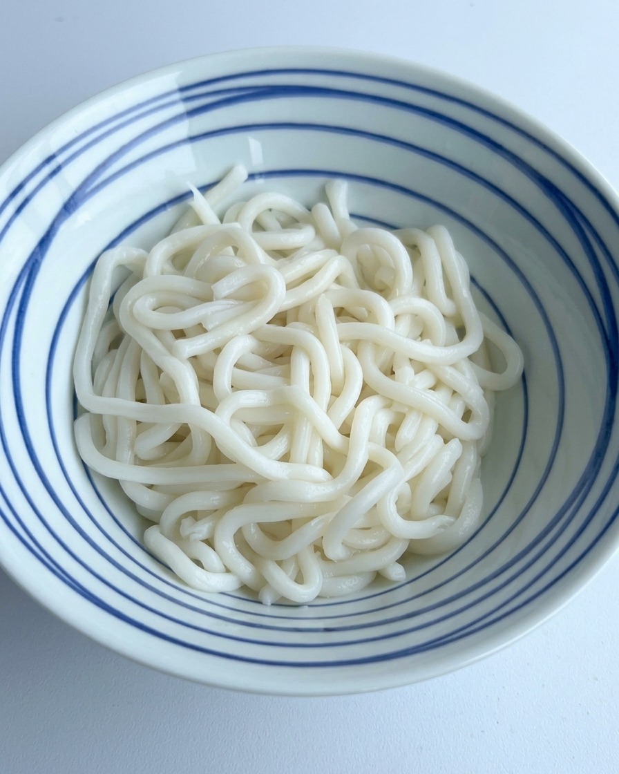 青いストライプの器に入った白いおうどんの上に、濃口醤油と調味料を回しかけている様子。