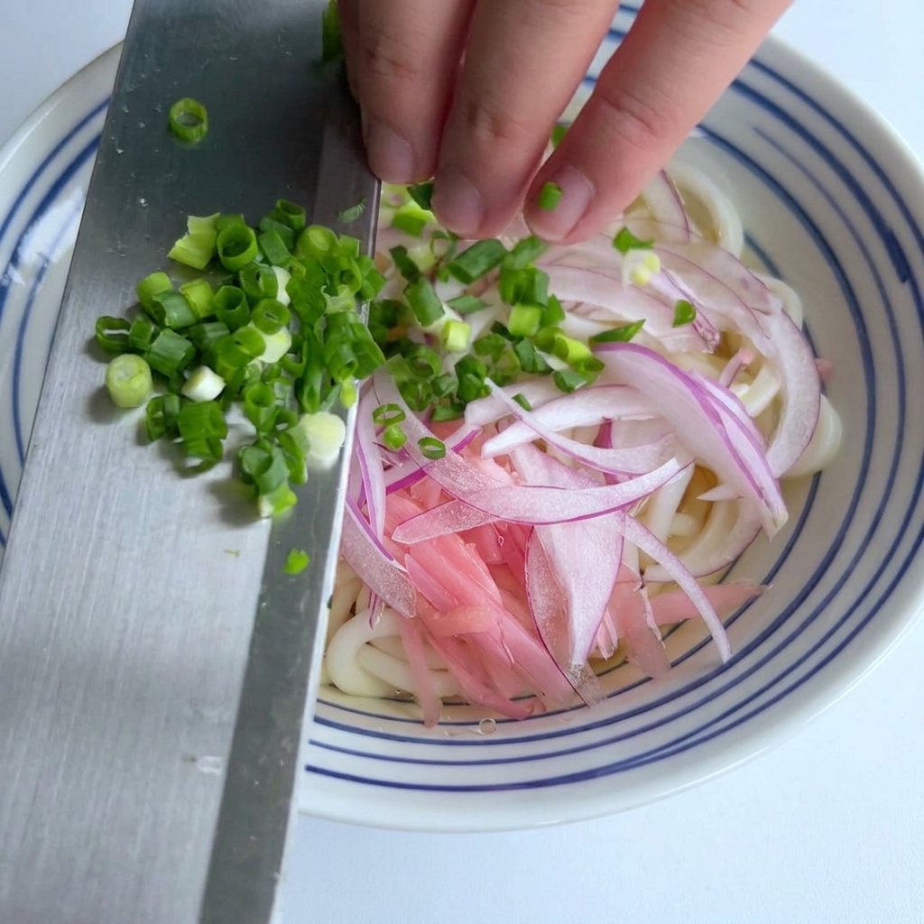 赤玉ねぎがのったうどんの器へ、包丁の側面を使って細かく刻んだ青ねぎをスライドさせて加える様子。