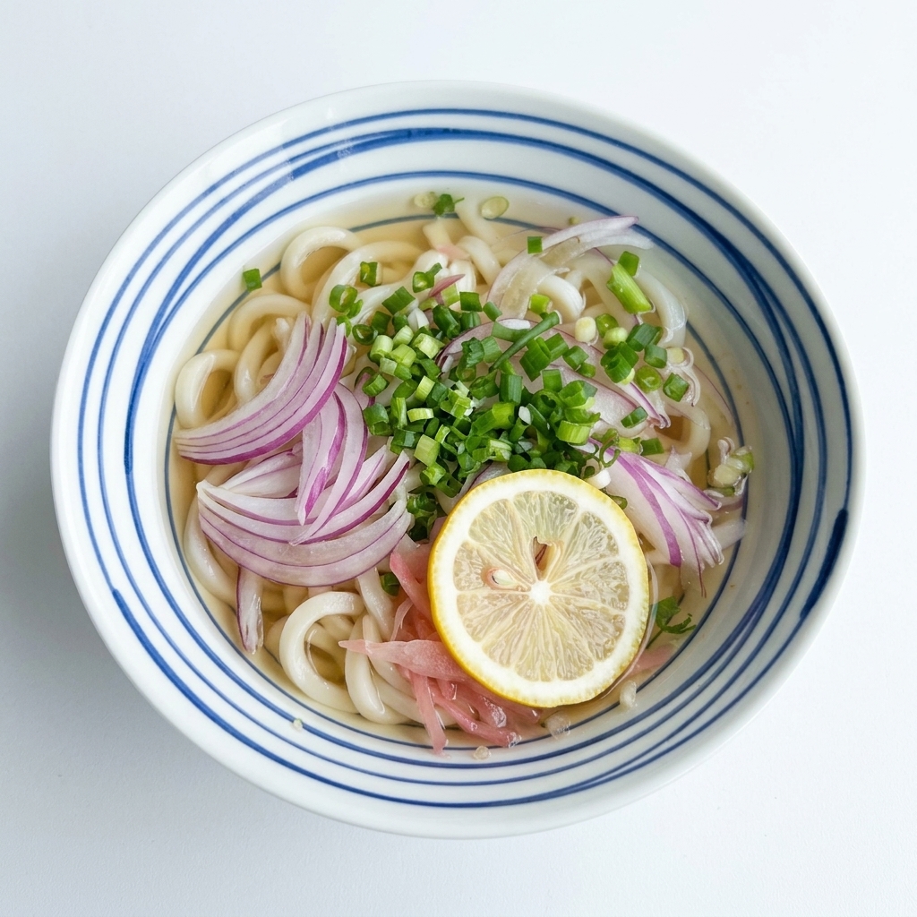 透き通ったつゆに浸かったうどんの上に赤玉ねぎやネギがのり、その中央に1枚の黄色いレモンスライスが配置された器の俯瞰写真。