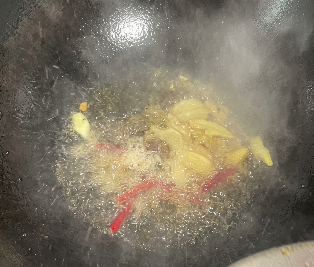 黒い中華鍋の中の熱い油で生姜のスライスと乾燥赤唐辛子が泡立ち、湯気とともに香気が立ち上っている様子。
