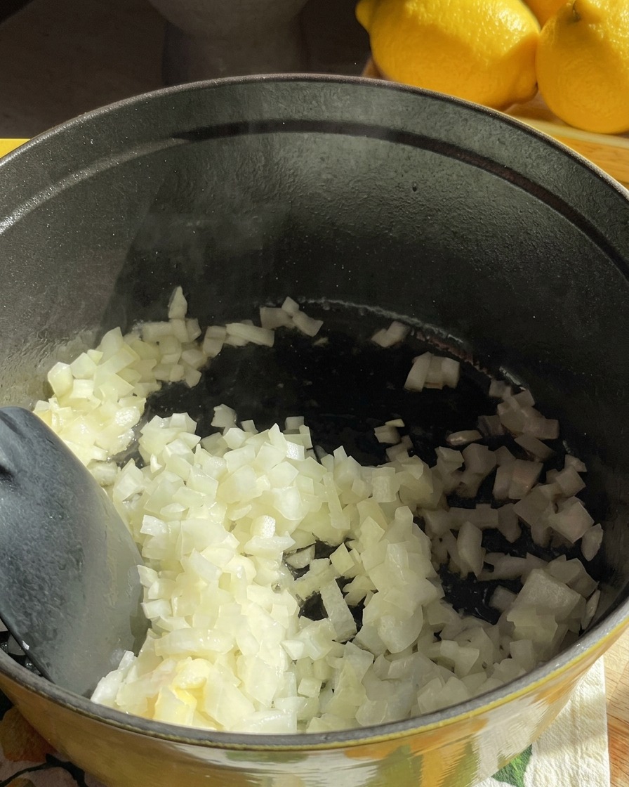 黒い調理鍋の中で、みじん切りの玉ねぎがバターでしんなりするまで炒められている様子。