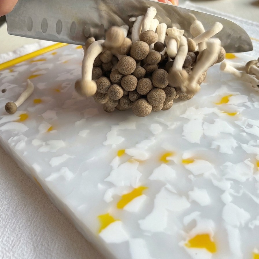 模様入りのまな板の上で、シェフナイフを使って茶色いしめじの株の根元を切り落としている様子。