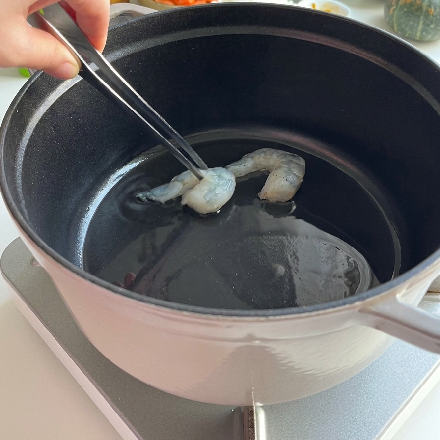 熱した油の入った黒い鍋に、金属製のトングを使って下味をつけた生エビを並べて焼いている様子。