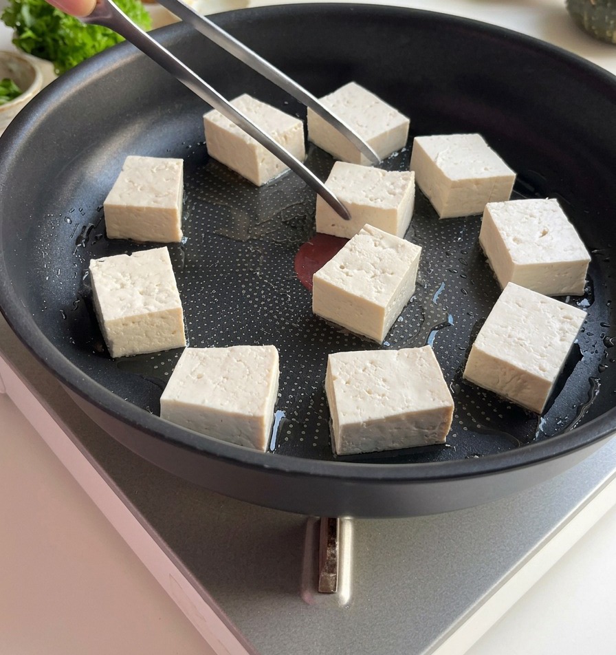 薄く油を引いた平らなフライパンに、トングを使って白い角切りの豆腐を並べている様子。