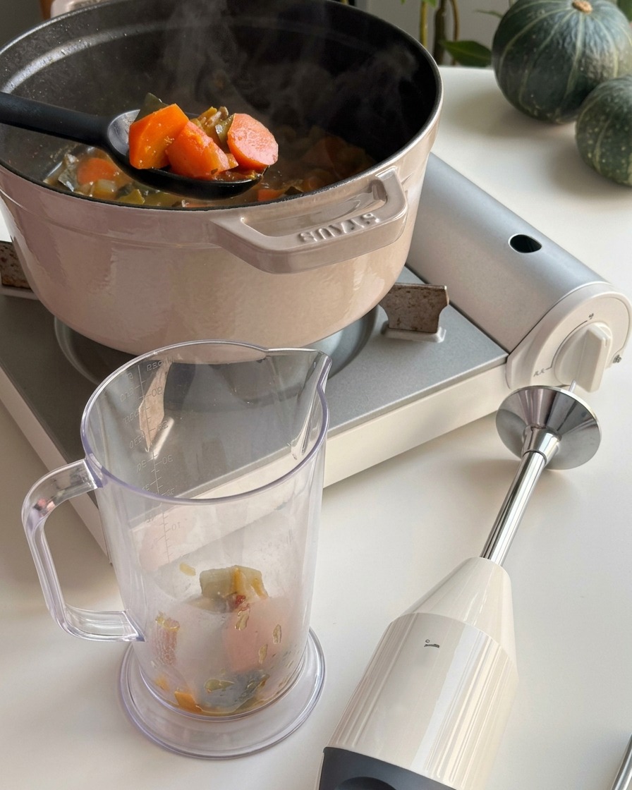 黒いレードルを使い、鍋からオレンジ色のかぼちゃの塊とスープをすくい、透明なプラスチック製のミキサー容器に移している様子。