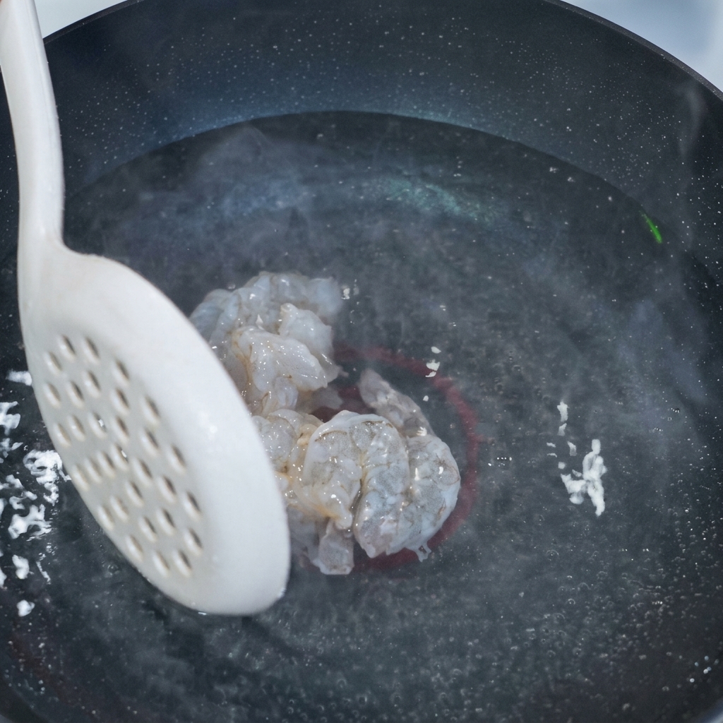 湯気が立つ鍋の上で、下味をつけた透明感のあるエビを数個すくい上げている白い穴あきスプーン。