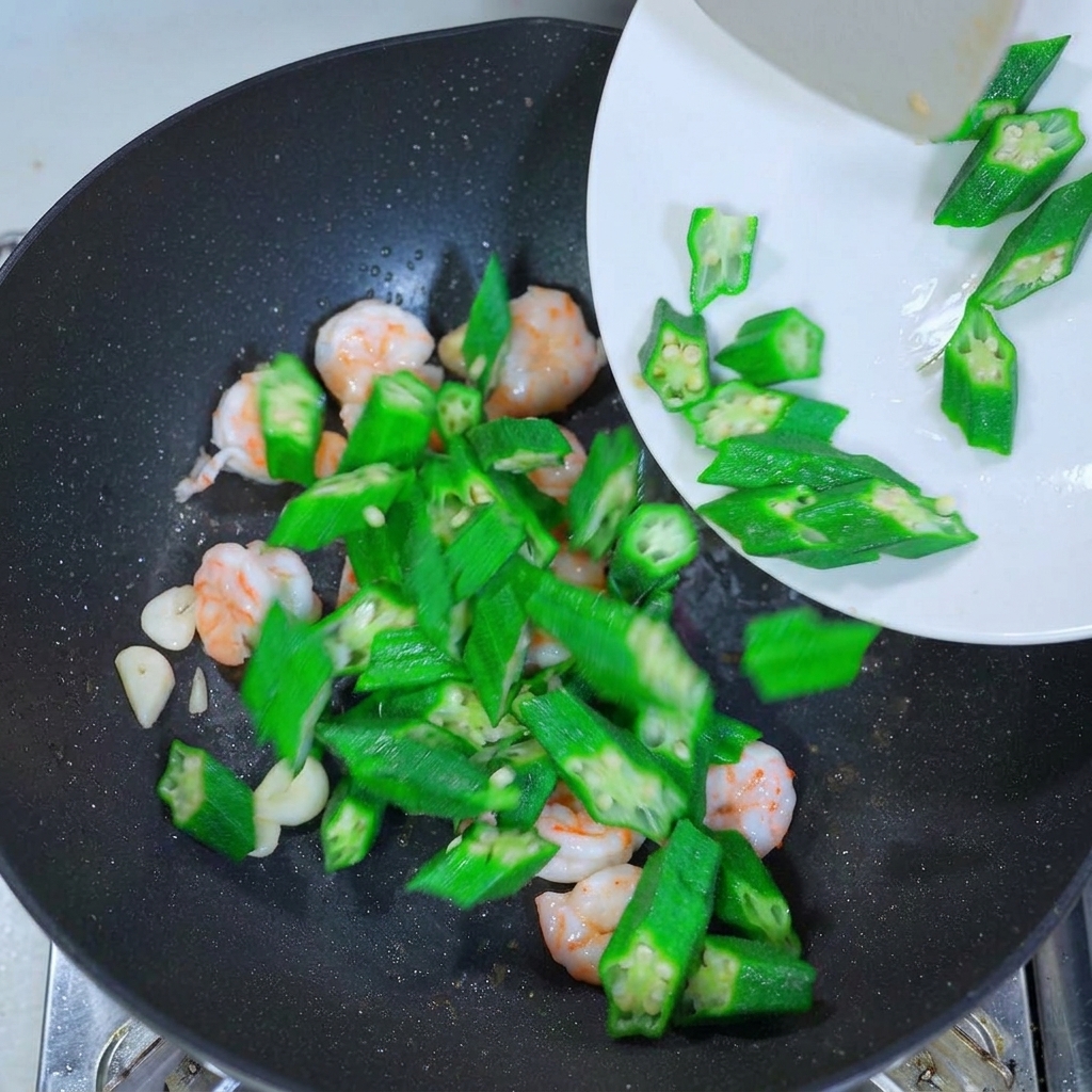 白いお皿から、炒めたエビとにんにくが入った中華鍋に、鮮やかな緑色のオクラが投入されている様子。