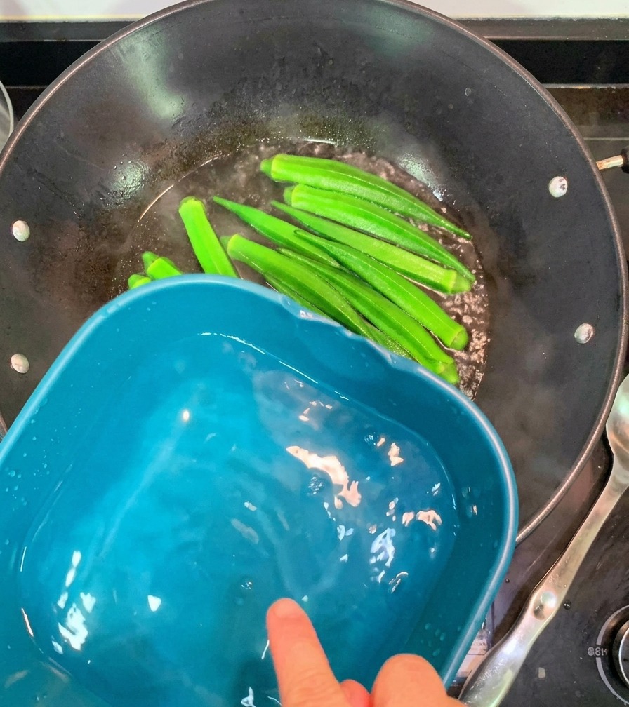湯気が立つ中華鍋から、鮮やかな緑色に茹で上がったオクラをすくい上げ、隣の冷水の入ったボウルに移している様子。