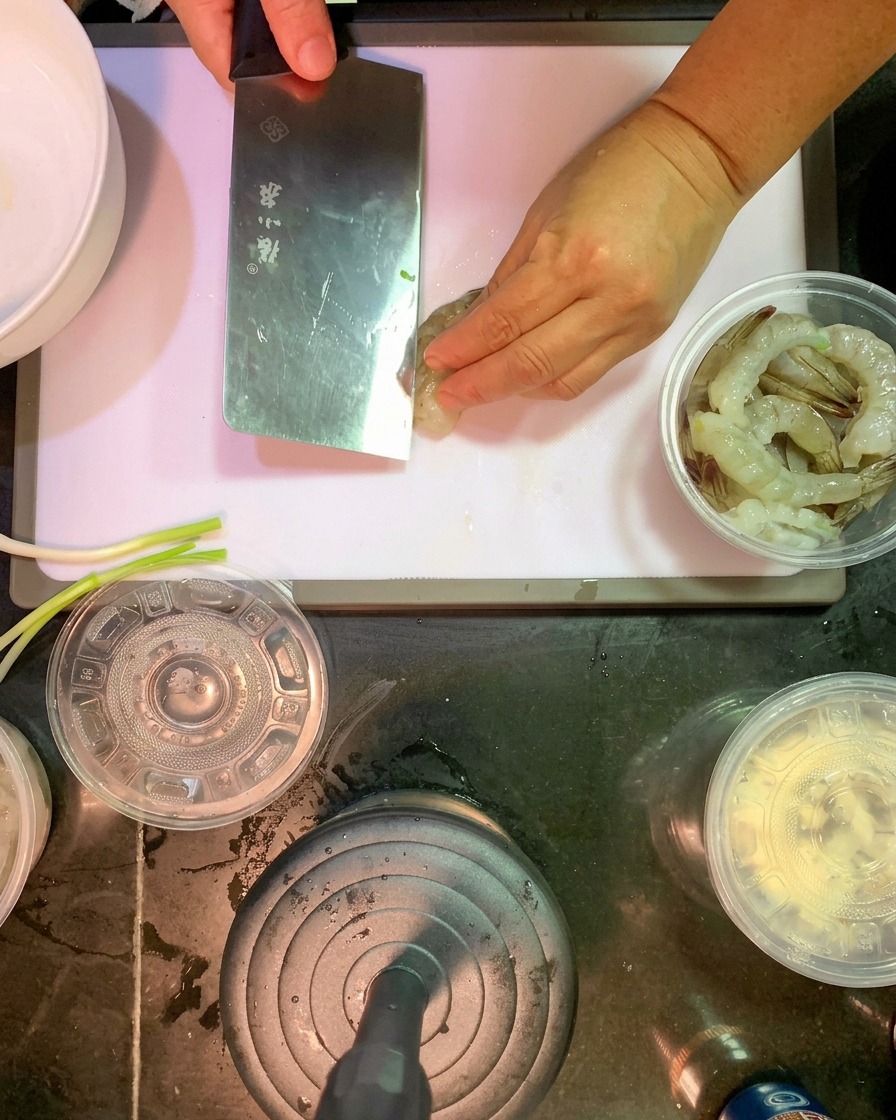 白のプラスチック製まな板の上で、殻をむいた生の海老の背に沿って、包丁で浅い切れ目を入れている様子。
