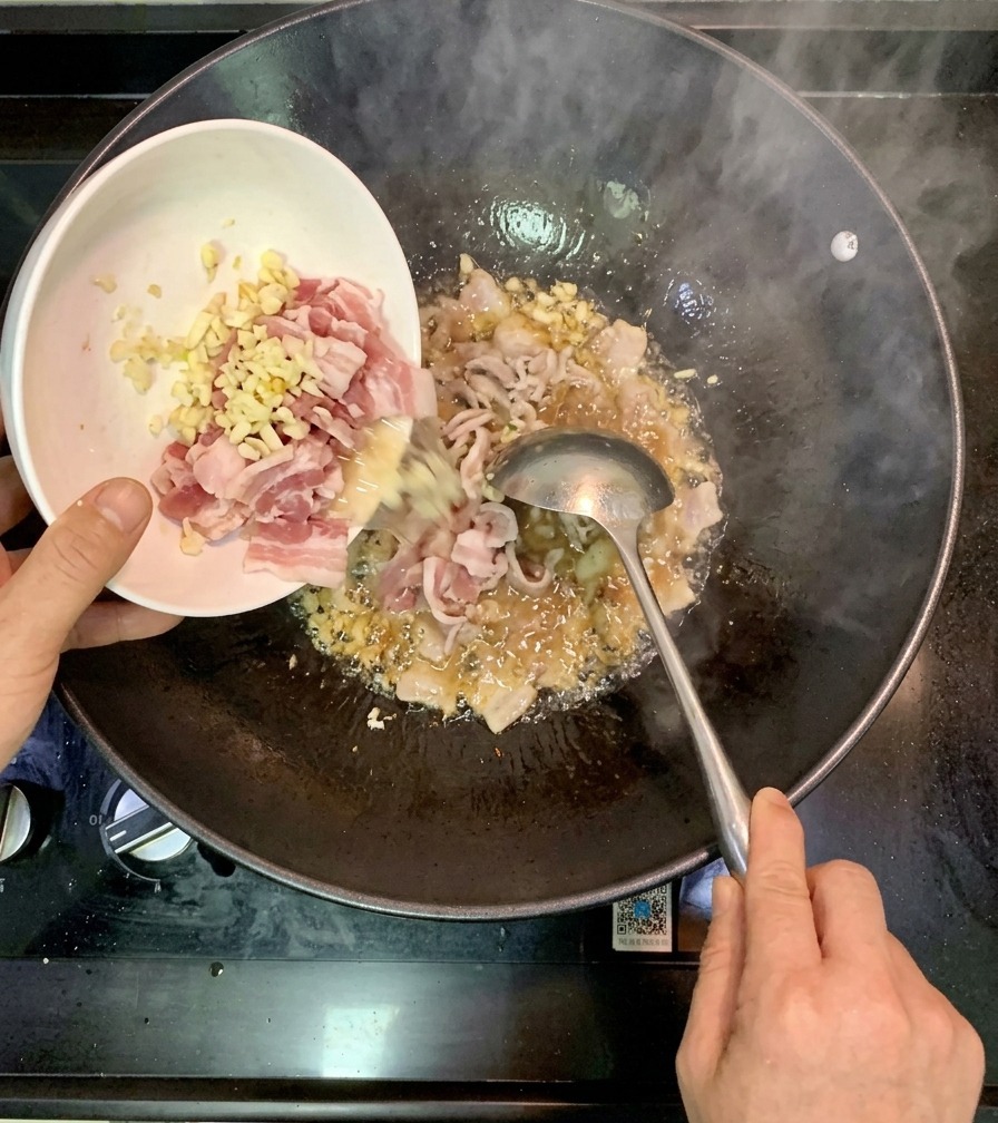 にんにくを炒めた熱い中華鍋の中に、小さな白いボウルから下味をつけた生の海老を投入している様子。