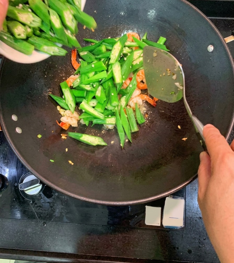 すでにピンク色に炒まったエビと薬味が入った中華鍋の中に、器から斜め切りにした緑のオクラを一気に投入している様子。
