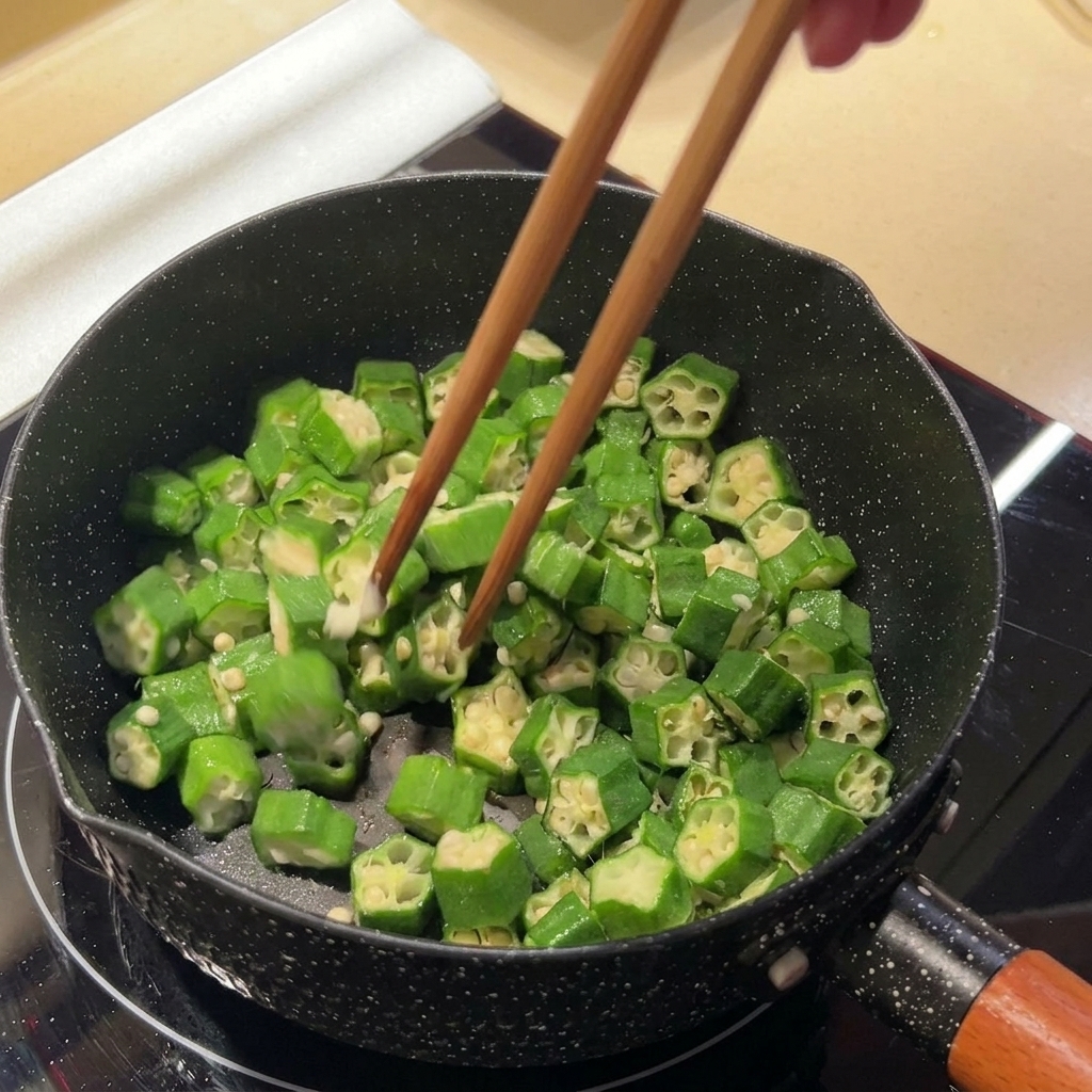 IHヒーターの上のフライパンで、菜箸を使ってスライスしたオクラを炒めている様子。