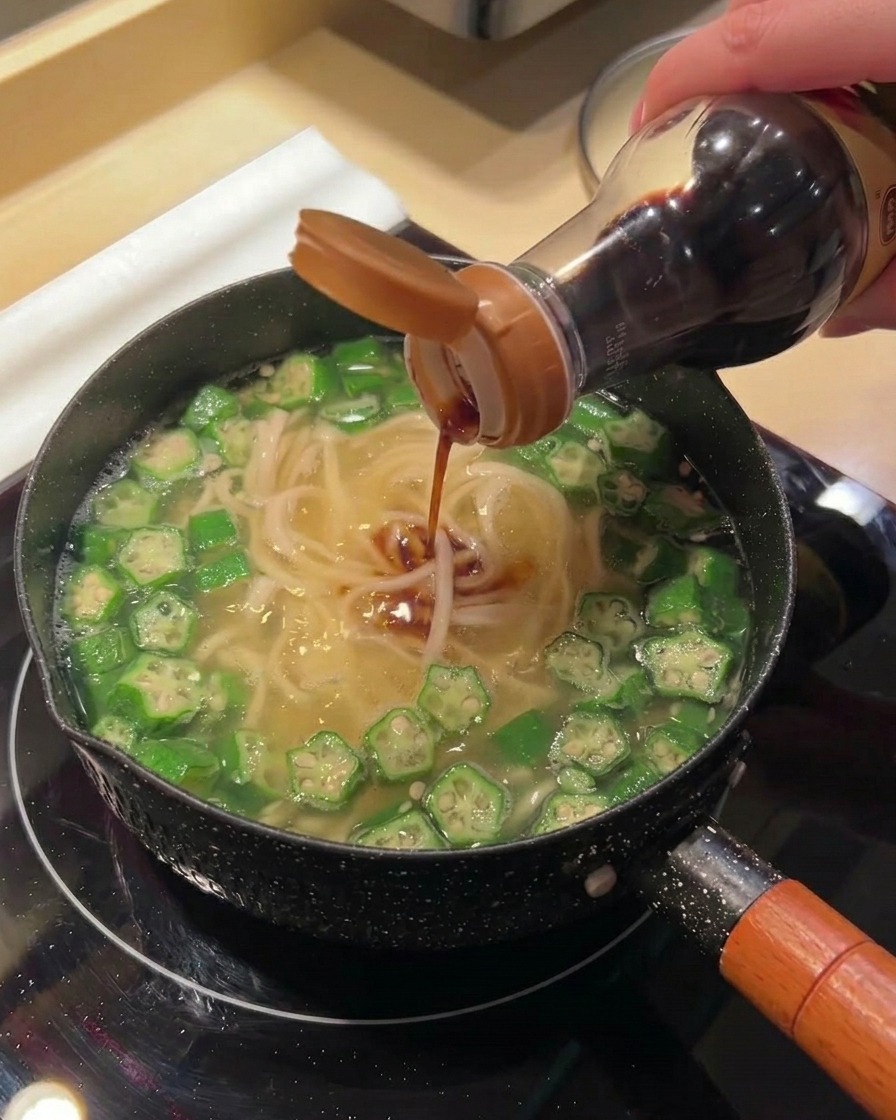 グツグツ煮立つ麺とオクラの鍋に、ボトルから醤油などの調味料を注いでいる様子。