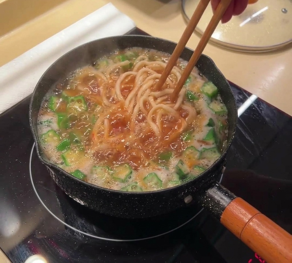 赤みを帯びた沸騰したスープの中で、長い麺と緑のオクラを菜箸でかき混ぜている様子。