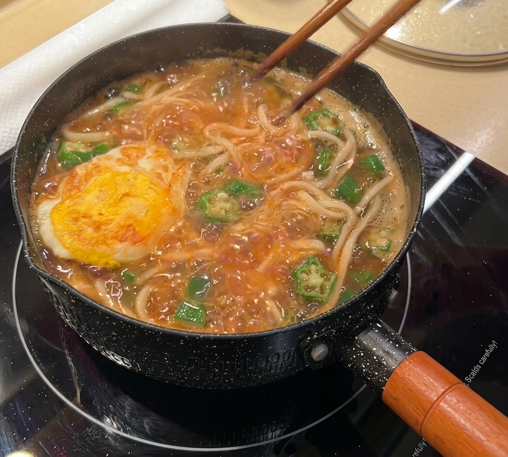 菜箸でこんがり焼けた目玉焼きを、煮立つスープ麺の上にのせている様子。