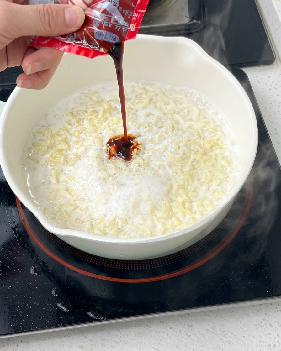 クリーミーな麺が入った白い鍋に、付属の調味料パックから真っ赤な激辛ブルダックソースを絞り出している様子。