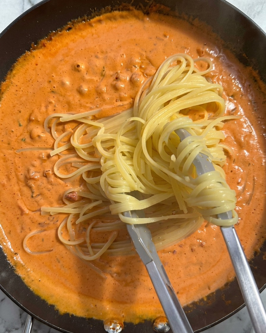 トングで茹で上がったパスタを掴み、濃厚なオレンジ色のクリームソースが満ちたフライパンへ移している様子。