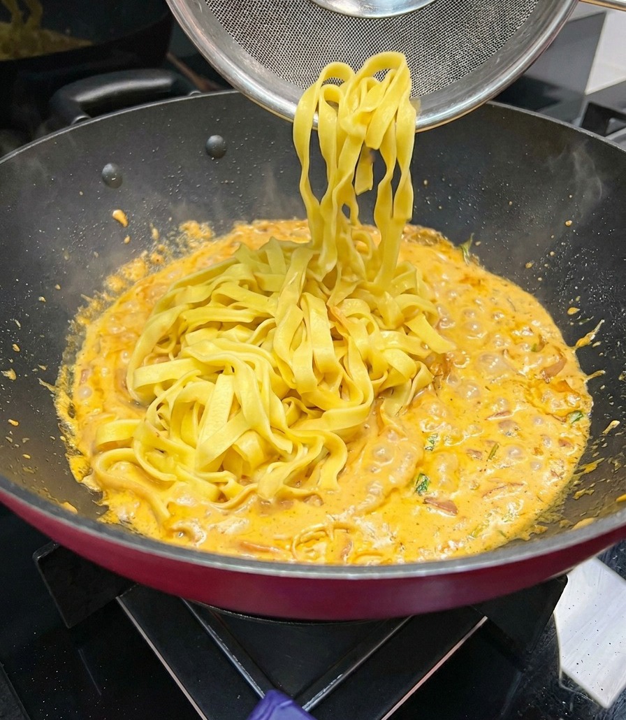 幅広のフェットチーネの麺を網じゃくしですくい上げ、フツフツと煮立つオレンジ色のクリームソースのフライパンに加えている様子。