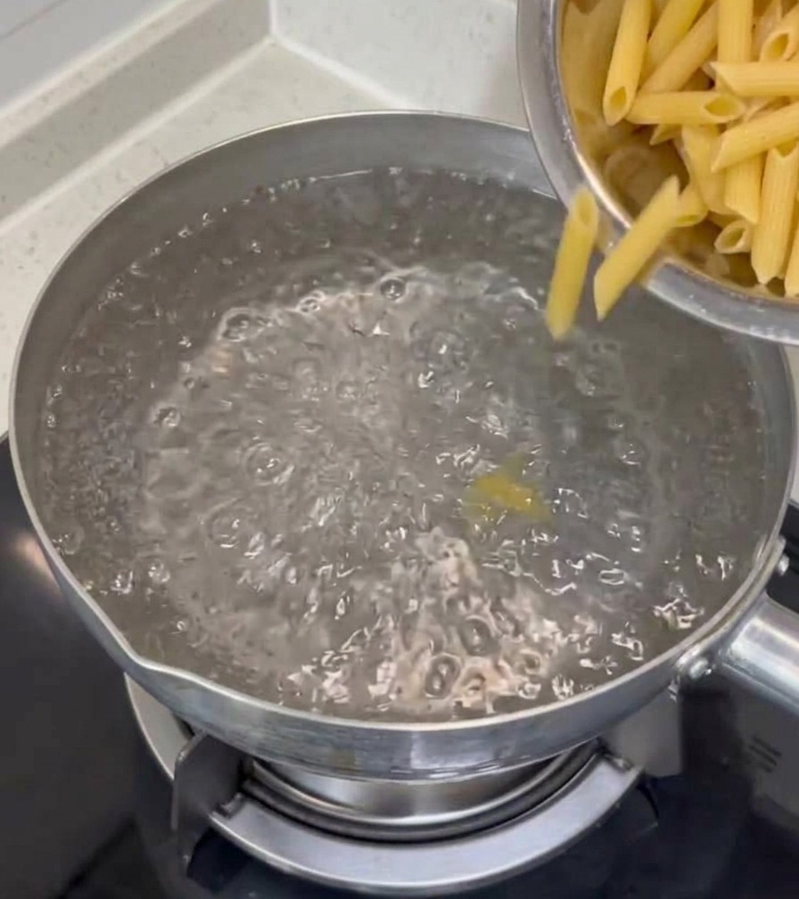 コンロの上の鍋で沸騰したお湯に、ステンレスのボウルから乾燥ペンネを入れている様子。