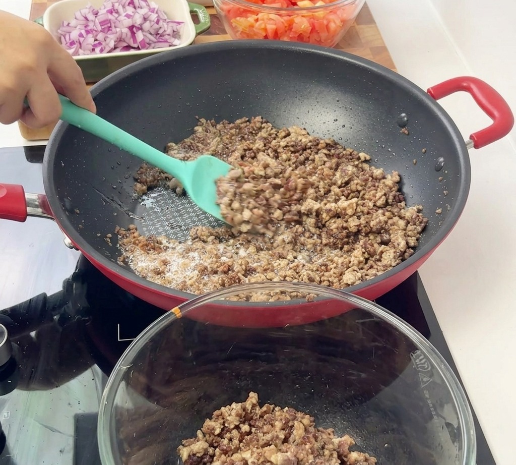 ティール色のスパチュラを使い、赤いフライパンから火の通った茶色い挽き肉をガラスボウルへと移している様子。
