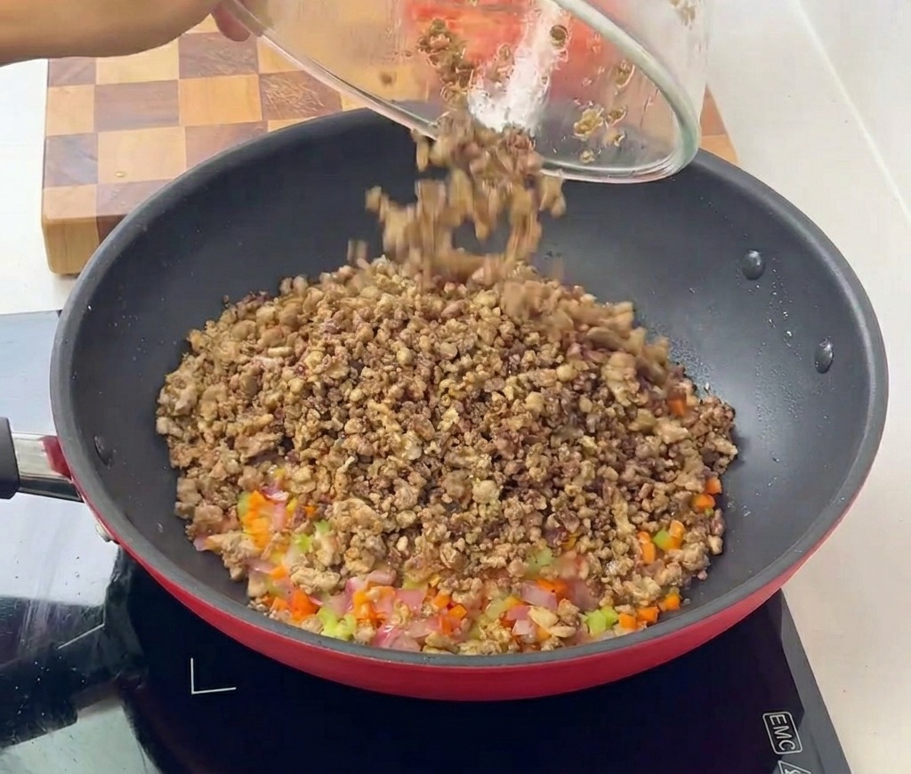 炒めたみじん切りの野菜が入ったフライパンに、ガラスボウルから火の通った挽き肉を戻し入れている様子。