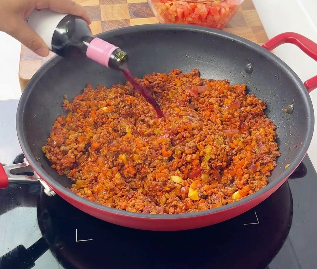 ボロネーゼの食材が入った赤いフライパンに、ボトルから赤ワインを注ぎ入れている様子。