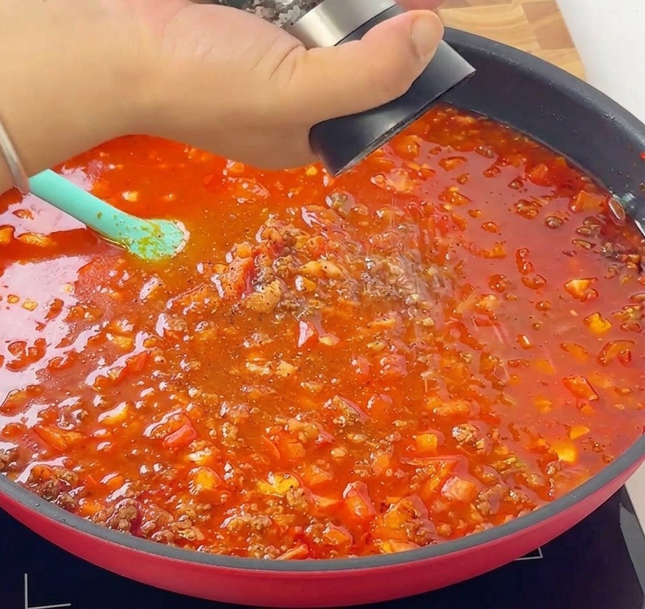 コトコトと煮立つお肉とトマトのソースが入ったフライパンの上で、ペッパーミルを手に持ち、挽きたての黒コショウを振りかけている様子。