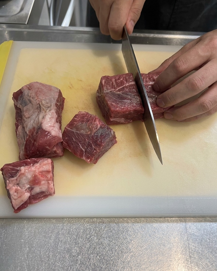 白いまな板の上で、きれいなサシの入った生の牛肩バラ肉を大きめの角切りにカットしている様子