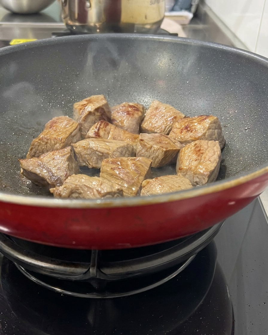 熱い赤いフライパンの中で、表面がこんがりときつね色に焼け、軽い湯気が上がっている角切りの牛肉