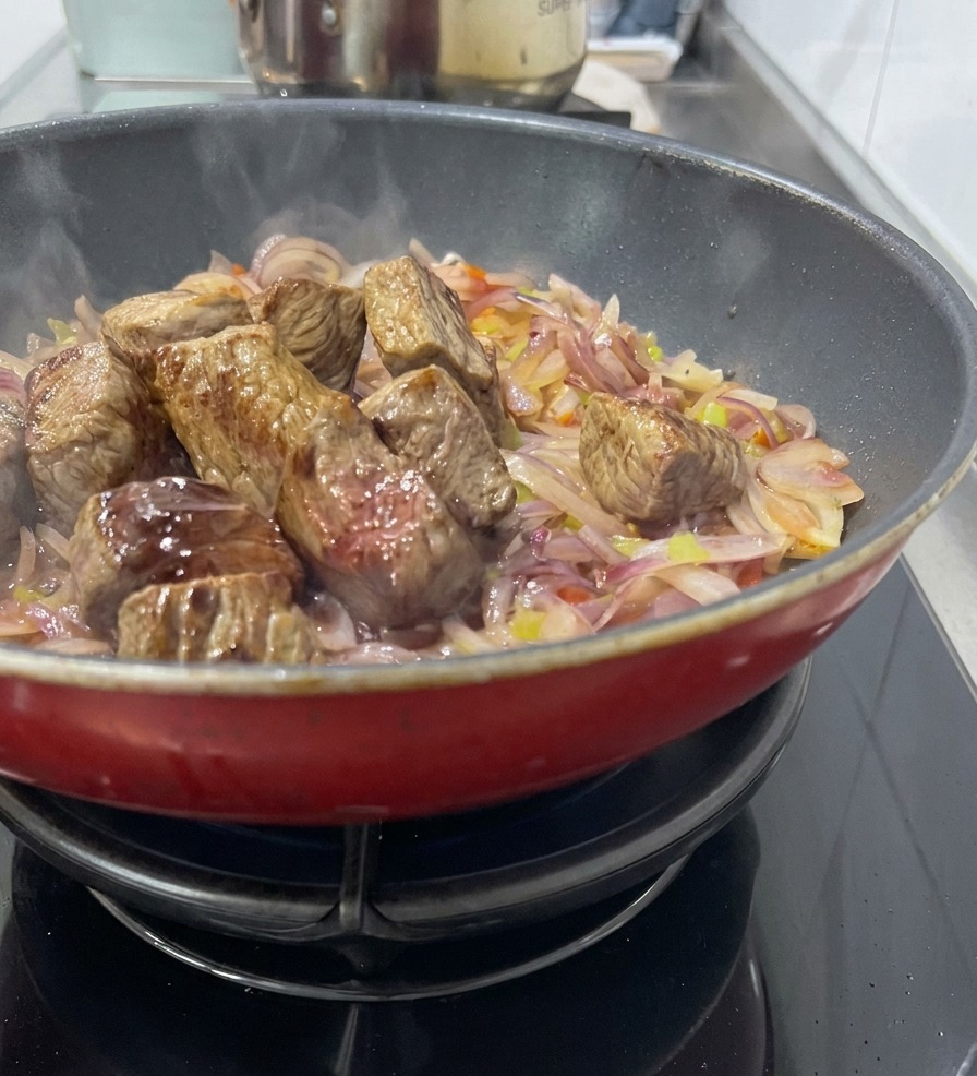 赤いフライパンの中で、しんなりした玉ねぎやセロリのベースの上にこんがり焼けた牛肉を戻し、赤ワインを注いで旨味を溶かし出している様子