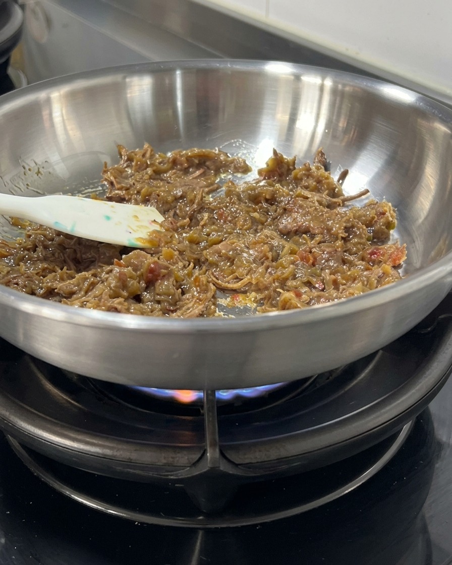ステンレスのフライパンの中で、濃厚なダークカラーのミートソースとほぐした牛肉を、ヘラを使って弱火で優しく混ぜ合わせながら温めている様子