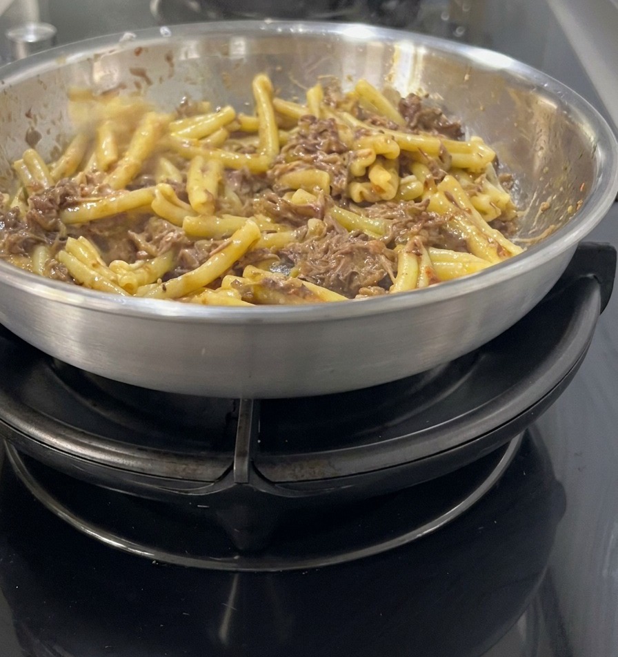ステンレスのフライパンの中で、茹で上がったカサレッチェパスタを濃厚な肉と玉ねぎのダークソースに優しく和えている様子