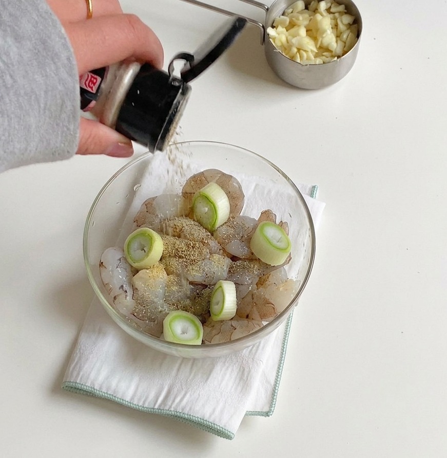 ガラスのボウルに入った生エビと刻んだ青ネギに、調味料の容器から塩とコショウを振りかけている手元