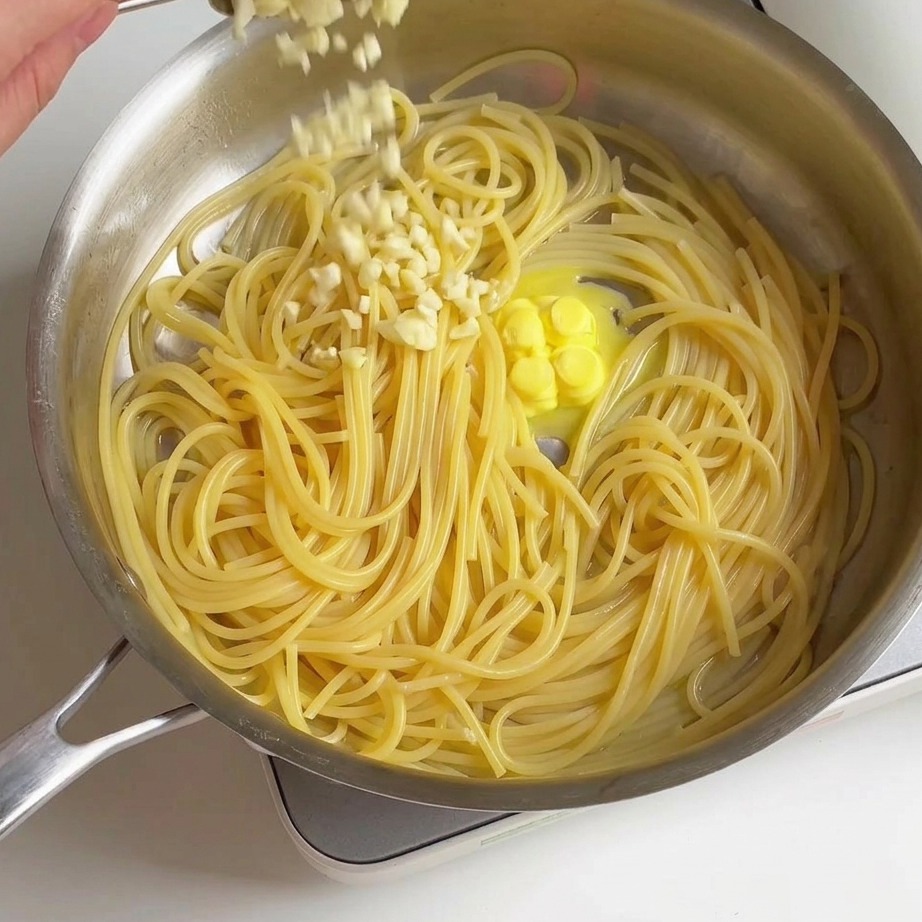 鍋の中の茹で立てのスパゲッティと溶けかけたバターの上に、スプーンから新鮮なみじん切りニンニクを散らしている様子