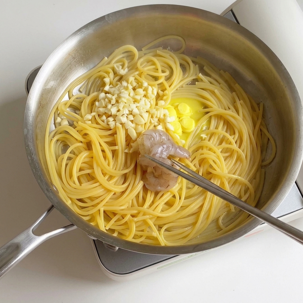 茹でたスパゲッティ、溶けたバター、みじん切りニンニクが入ったステンレス製の鍋に、マリネしておいたエビを投入している様子