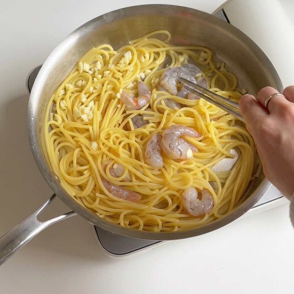 フライパンの中でエビとスパゲッティを炒め合わせており、エビがだんだんとピンク色で不透明に変わっていく様子