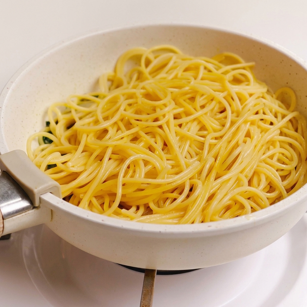 フライパンの中で、緑のハーブ、にんにく、ベーコンのソースの上に、茹で上がったスパゲッティが載せられている様子。