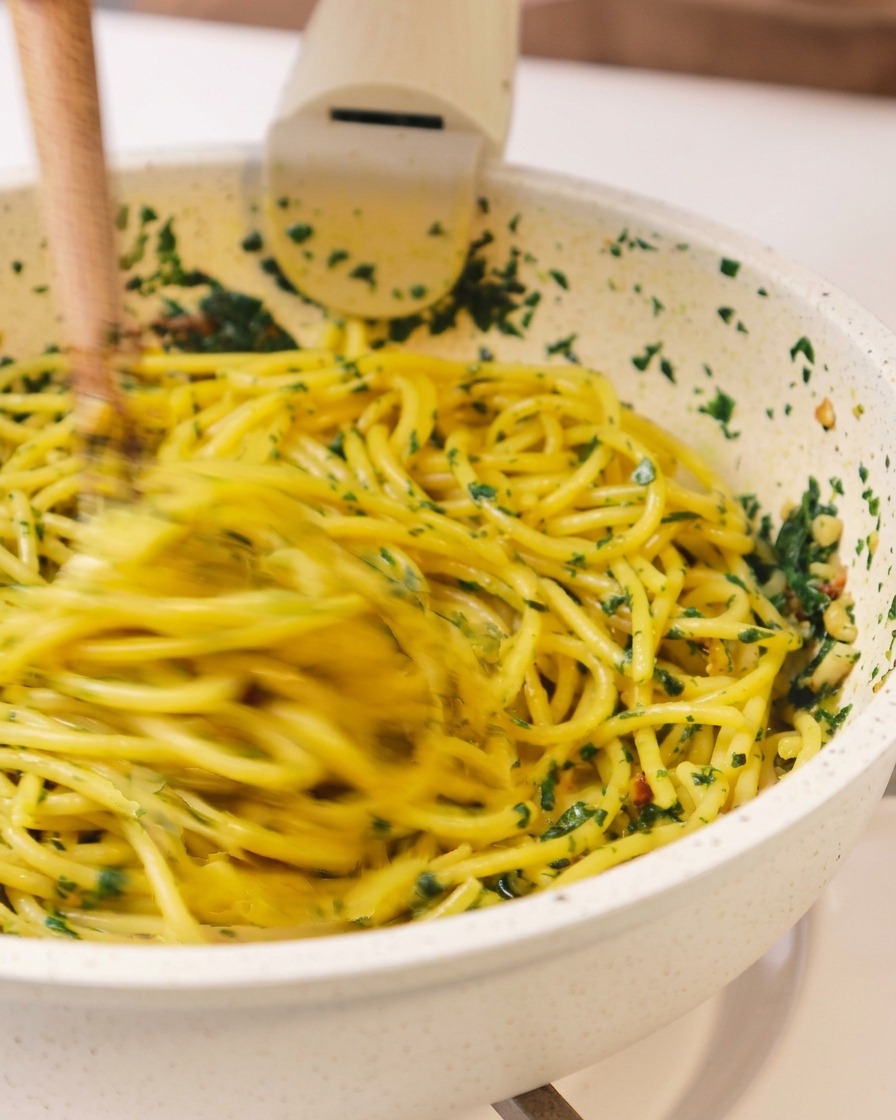 フライパンの中で、木箸を使い、緑のハーブとオイルのソースをスパゲッティと勢いよく混ぜ合わせている躍動感のあるカット。