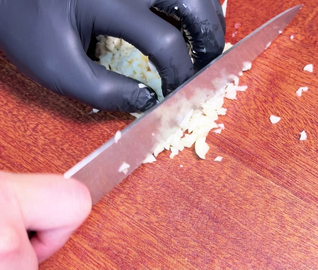 黒い手袋を着用した手が、暗い色の木製まな板の上で、大きなシェフナイフを使って生のニンニクのクローブを細かくみじん切りにしている様子。