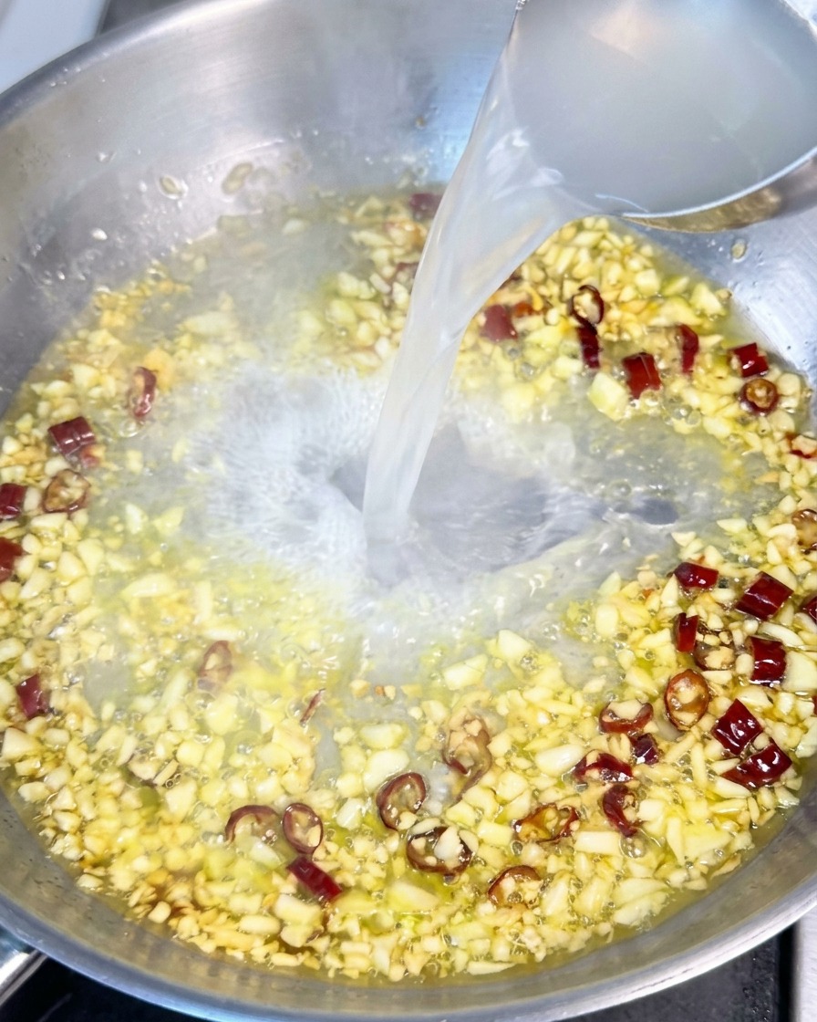 おたまを使って、にんにくと赤唐辛子が入ったフライパンに、白く濁ったパスタの茹で汁を注いでいる様子。