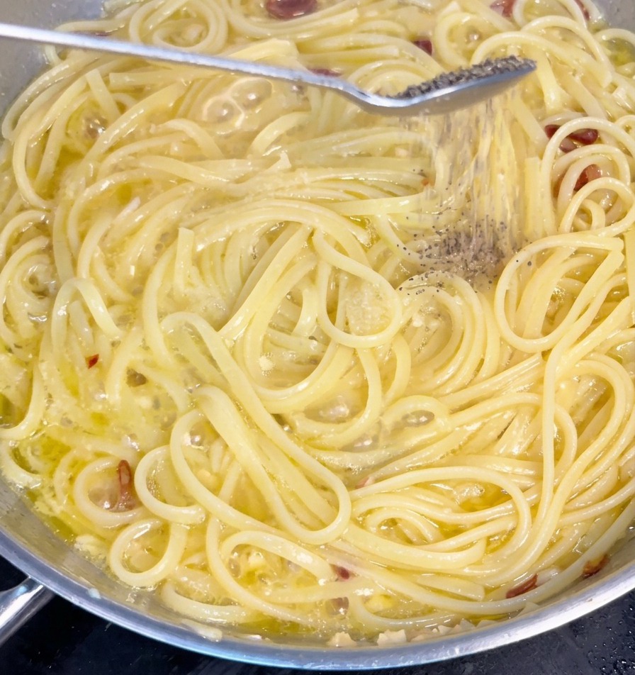 にんにくオイルソースで軽く煮立っているスパゲッティの麺の上に、スプーンから挽きたての黒コショウが振りかけられている様子。