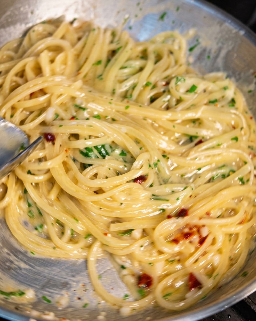 スパゲッティが入ったステンレス製のフライパンをトングで激しく煽っている様子。にんにくのみじん切り、赤唐辛子、緑のパセリが混ざり合い、麺全体がツヤのあるクリーミーなソースで綺麗にコーティングされている。