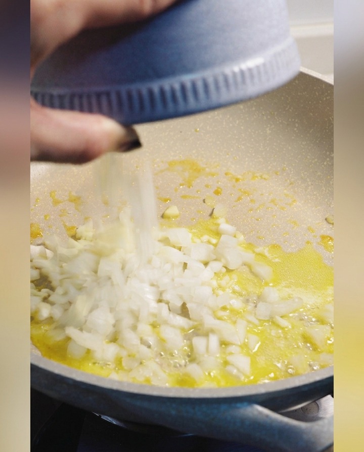 溶けたバターが入ったフライパンに、青いボウルからみじん切りにした白い玉ねぎを加えている様子。