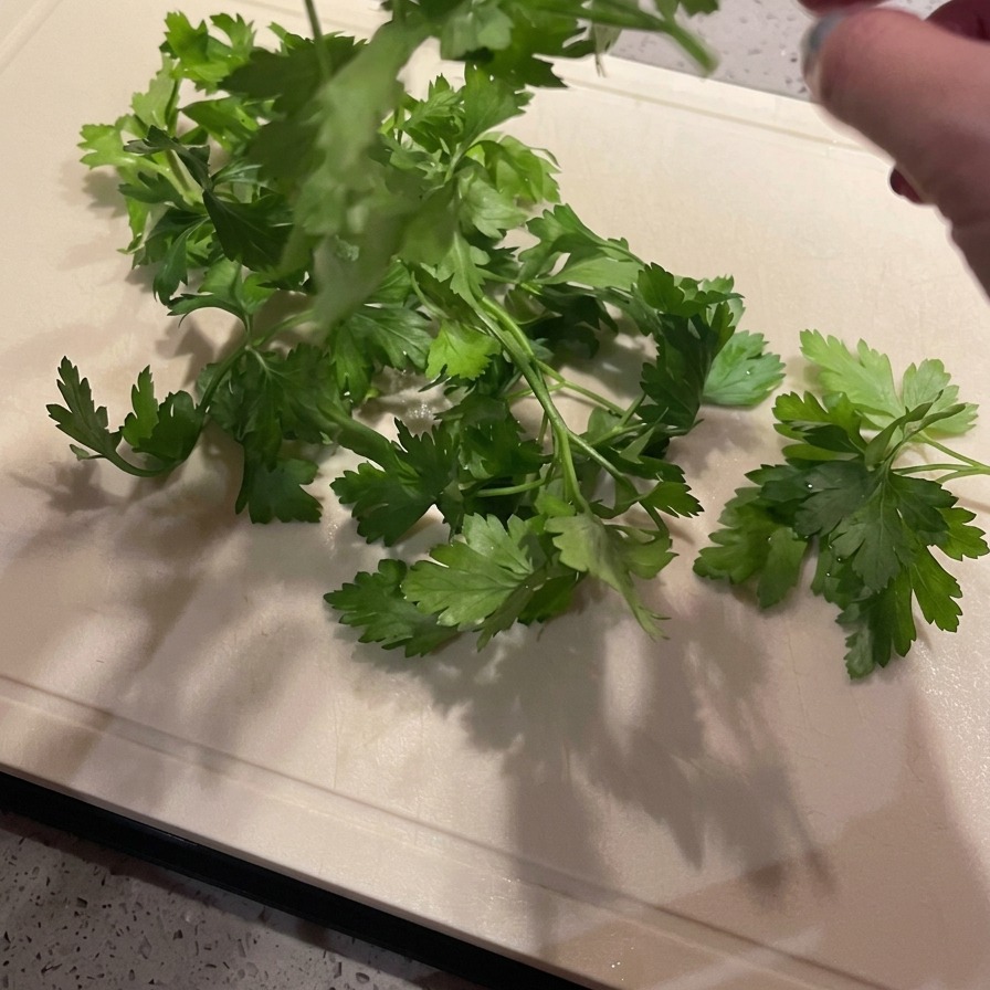 明るい色のまな板の上で、鮮やかな緑色のイタリアンパセリの茎から葉だけを丁寧に手で摘み取っている様子。