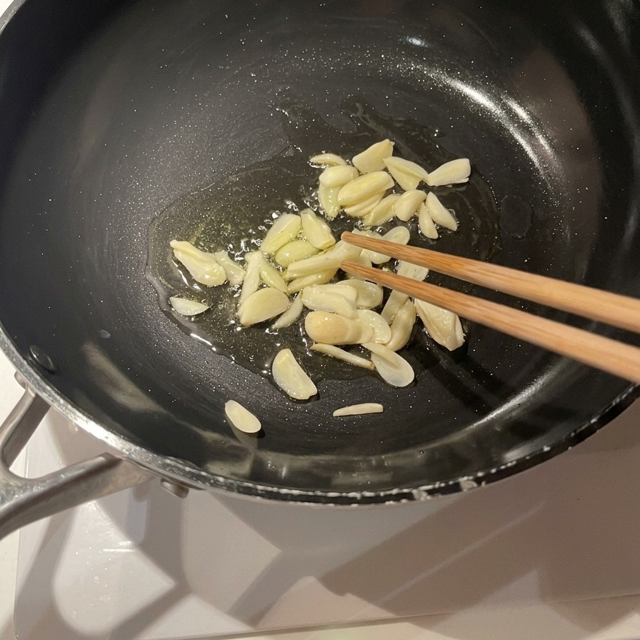 黒いフライパンに張られたオリーブオイルの中で、スライスしたにんにくがシュワシュワと泡立ち、木箸で混ぜられている様子。