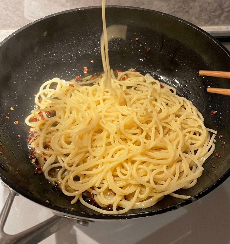 茹であがった長いスパゲッティの麺を、金属製の網じゃくしで茹で汁から直接すくい上げ、にんにくオイルの入ったフライパンへ移している様子。