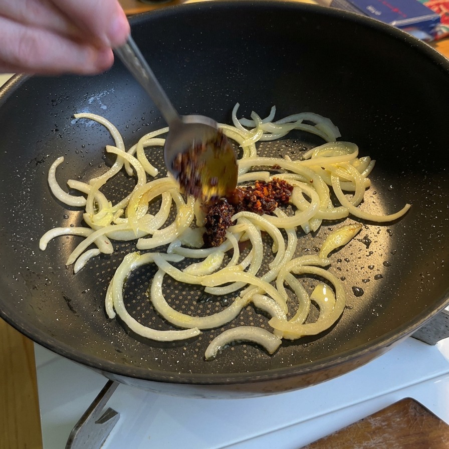 スライスした玉ねぎが炒められているフライパンに、スプーンで真っ赤なチリオイルを加えている様子。