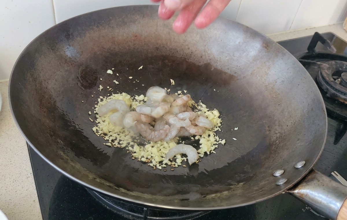 にんにくを炒めている熱い中華鍋の中に、生の透明感があるむき海老をひとつかみ投入する様子。