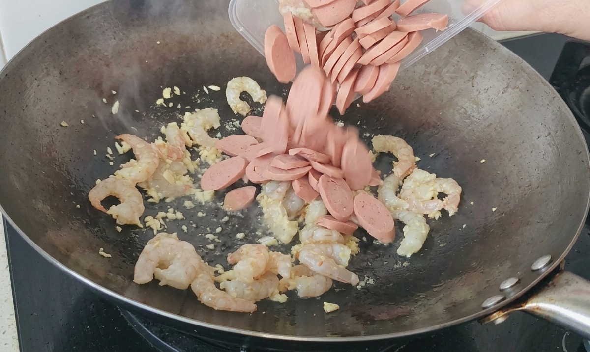 海老とにんにくを炒めている中華鍋に、プラスチック容器からスライスしたソーセージを投入している様子。