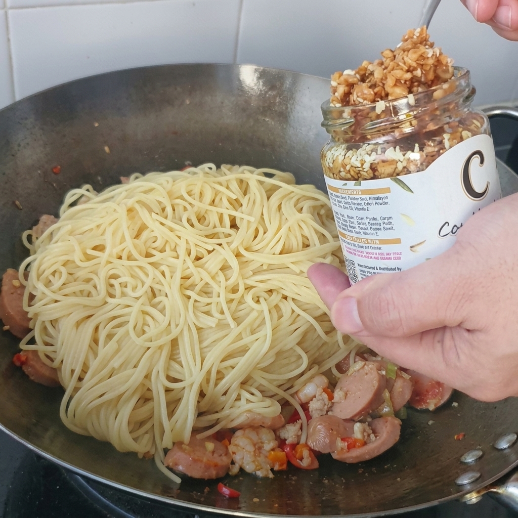 金属製の中華鍋に入ったスパゲティの山の上に、ガラス瓶からスプーンでサクサクのフライドガーリックをすくい乗せる様子。